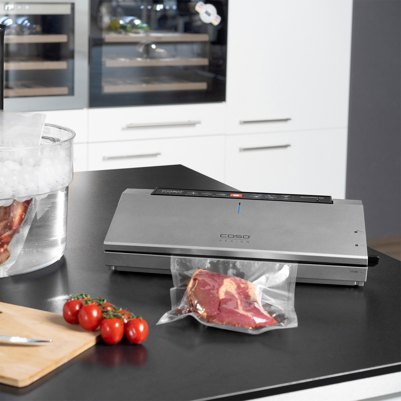 Vakuumiergerät mit Fleisch und Tomaten auf einer schwarzen Oberfläche in der Küche.
