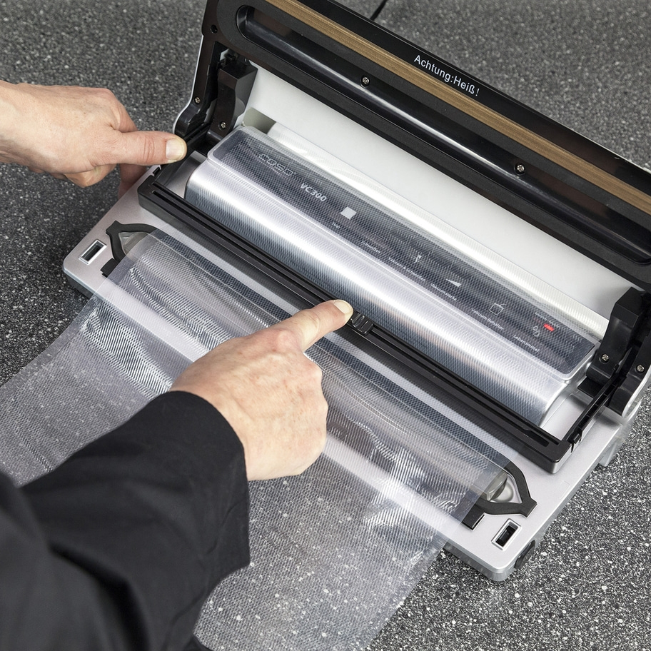 Person bedient Vakuumiergerät. Zeigt auf Folie. Achtung: Heiß! Coso VC300.